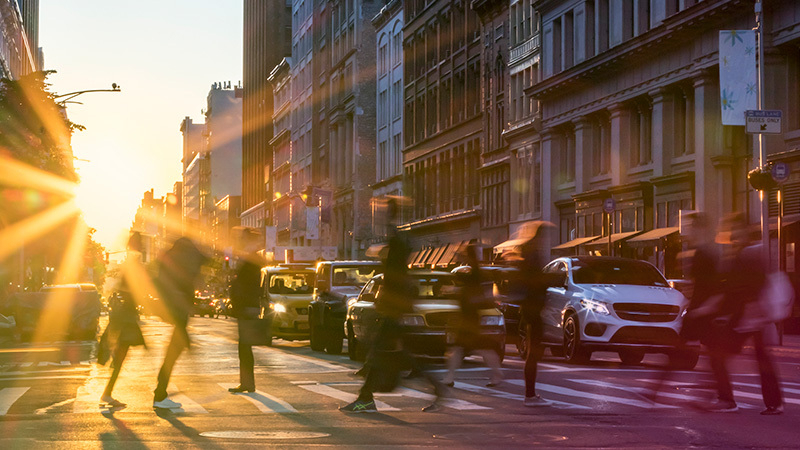 Busy city street with people crossing at an intersection, sun setting in the background